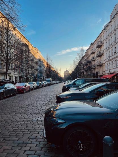 View down an avenue with parked cars and old buildings. The top floor of the buildings on the left is illuminated by the setting sun. The street ends with a view of the Berlin TV tower and the water tower next to it, somewhat diffused in the sunlight.