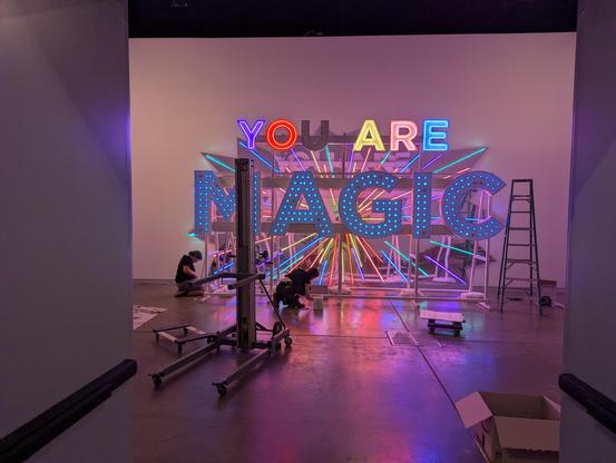 Big sign sculpture framed by a door with the U out in YOU and a material lift and ladder and people working on the sculpture in the shot. The room is multicolor with a shade of pink and the MAGIC letters illuminated by blue light bulbs