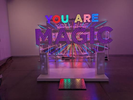 Your Magic Is Real sculpture all lit with the U remade, purple MAGIC and pedestal in place, YOU ARE letters reflecting off of the metal ramp leading to the pedestals and the room glowing a warm pink