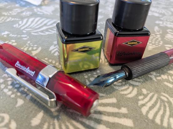 A red Kaweco sport and a dip pen in front of a pair of Diamine 12 ml bottles, one gold, one red. On a gree tablecloth with shell motifs.