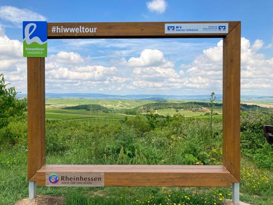 Ausblick auf eine grüne, leicht hügelige Landschaft. Umrahmt von einem Holzrahmen, auf dem oben links das Logo der sog. Hiwweltouren mit dem Wort „Heideblick“ steht. Daneben #Hiwweltour. Rechts oben am Rahmen klebt Werbung, unten links ein Schild „Rheinhessen“. Der Rahmen ist nicht die Grenze des Bildes, rundherum sieht man auch die Landschaft, quasi als 2. Rahmen.