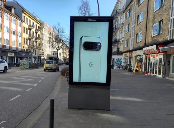 Massiver Bildschirm mit Google-Konsumpropaganda auf dem Gehweg direkt neben dem gestrichelten Radweg.