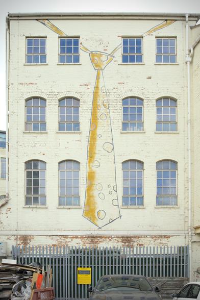 The side of an old building. It has three floors of windows. It's painted a light yellow, maybe a custard yellow, but the paintwork is peeling. A tie has been painted in the centre - as if the building is wearing a tie. The tie has a grey outline, with grey dots and has a deeper, mustard yellow highlighting one side. At the bottom of the building is a metal fence. The tops of cars and a skip can be seen this side of the fence.