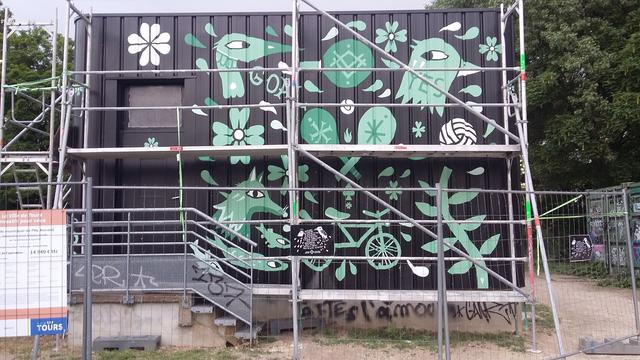 Chantier avec échafaudages de peinture verte soir fond noir. Motifs divers et fantaisistes visant à symboliser l'activité sportive sur un bâtiment qui sert de vestiaire. Au bas est tagué  : "faites l'amour"