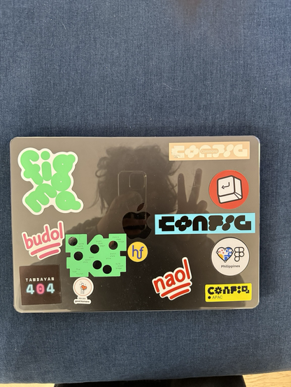 Top-down shot of Chi’s MacBook with a clear case, reflecting her silhouette and peace sign in the glossy surface. The case is decorated with vibrant stickers from Figma Config 2025, including 'Config Commons,' 'Config APAC,' 'Philippines Friends of Figma,' and more. Other stickers include Tambayan 404, Humanforce, Next Keyboard Club, and fun phrases like 'budol' and 'naol.'