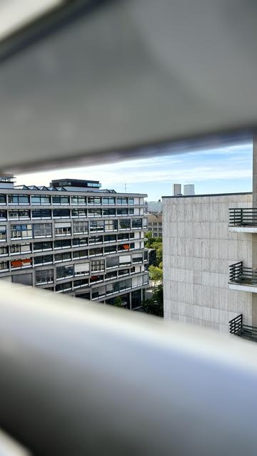 Auf dem Foto sind im Vordergrund unscharf die Lamellen von Fensterläden zu sehen. Im Fokus sind mehrere Gebäude (links das Architekturgebäude der TU Berlin). In der Mitte sind Bäume zu sehen sowie weitere Gebäude.