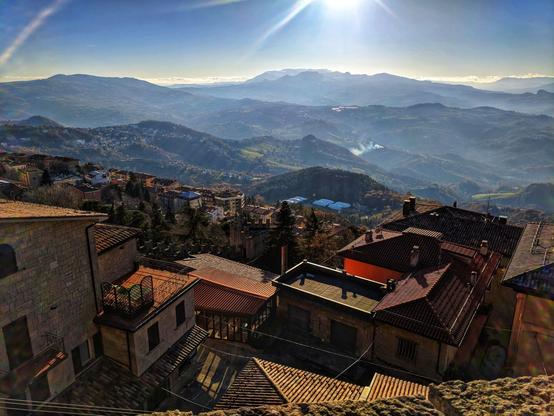 A scenic view of Guaita Tower and Monte Titano in San Marino. The image captures a mountainous landscape under a bright, sunny sky. The tower stands prominently amidst a cluster of buildings with red-tiled roofs. In the background, layers of hills fade into the distance, creating a sense of depth. The sunlight casts a warm glow over the scene, highlighting the textures of the buildings and the natural beauty of the surrounding landscape.