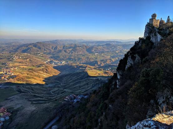 A scenic view of Guaita Tower atop Monte Titano in San Marino. The tower stands on a rocky cliff, overlooking a vast landscape of rolling hills and valleys under a clear blue sky. The foreground shows the rugged terrain leading up to the tower, while the background reveals the distant towns and the layers of hills fading into the horizon.