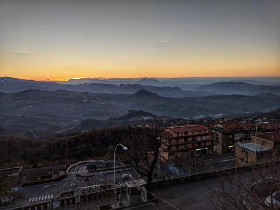 A scenic view of San Marino at sunset. The image captures a mountainous landscape with a vibrant sunset illuminating the horizon. Layers of hills fade into the distance, with a town visible in the foreground. The warm glow of the setting sun casts a golden light over the scene.