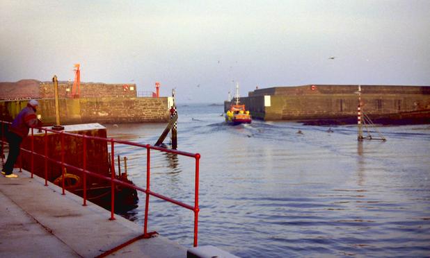 A harbour mouth, with a fishing boat departing. On the left, a man watches it, leaning on red railings.