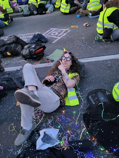 Ich liege auf der Straße und trage dabei ein buntes Hemd mit Blumenmustern, dazu eine Sternsonnenbrille mit Kulleraugenaufklebern drauf die nach links oben schielen, dazu mache ich mit meiner Hand eine Denkerpose. Ich trage einen Partyhut, eine gelbe Warnweste und bin mit bunten Luftschlangen und einer Menge konfettig überworfen. Um mich herrum sitzen Menschen auf der Straße und Rücksäcke sind zu sehen. Auf der Straße ist mit Kreide die Zahl161 gemalt.