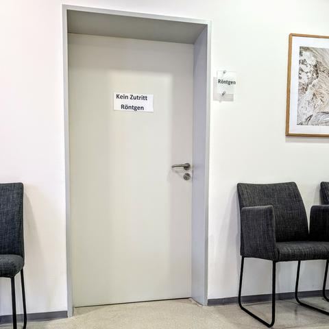 Weiße Tür mit der Aufschrift „Kein Zutritt – Röntgen“ in einer Arztpraxis. Links und rechts davon stehen graue Stühle im Wartebereich. Die Szene wirkt nüchtern und sachlich.
White door in a medical office marked “No entry – X-ray”. Grey chairs line the waiting area on both sides. The scene is minimal and clinical.
Foto by Johann Seidl, Gartenpoet.