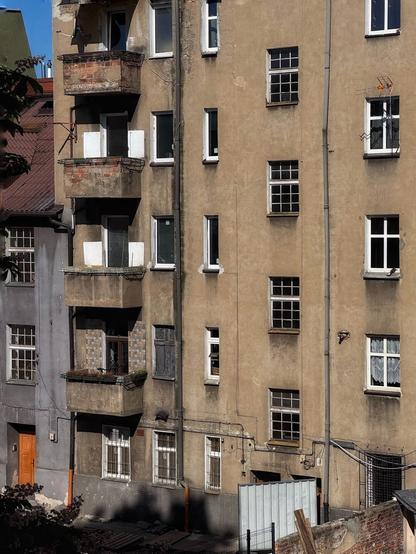 Ein verwittertes Wohnhaus mit mehreren Fenstern und Balkonen. Das Äußere ist größtenteils beige, wobei einige Bereiche Abnutzung und abblätternde Farbe zeigen. Verschiedene Texturen und Materialien sind sichtbar, darunter Ziegel-, Gips- und Metallelemente, die eine städtische Umgebung widerspiegeln.