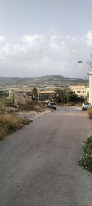 Looking down the hill from Lippija towards Gnejna and Mgarr in the distance. The land is dotted with shimmering silver poly tunnels and agriculture land in different shades of green, and tan, all the way up the hill that meets the horizon, and the blue hazey morning sky