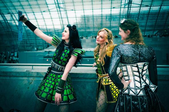 The photograph captures three young women in shiny cosplay outfits, posing as protagonists from the musical Six. The background reveals a modern architectural design with a prominent glass roof. In the foreground, the women are positioned by a metal railing, posing for a selfie.