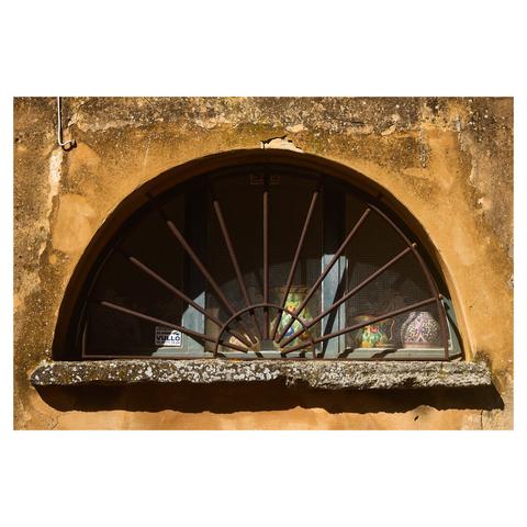 Flat image of a semicircular window in an ochre coloured, weathered facade. A metal grate protects the window. Behind the glass, colourful ceramic jugs are visible.