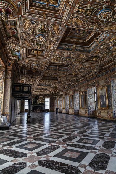 A portrait view at one corner of a large, ornate hall used for entertaining guests. It's empty of any furnishings giving a clear view of the tiled marble flooring in geometric patterned squares and diamonds of black, white, and dusky red colours. On the near left wall, halfway along the length of the hall, there is a a black lacquered balcony above head height supported by thin, black, lacquered columns. The ceiling is very busy with decoration, formed within a chequerboard pattern of recessed and flat squares on which there are brown lacquered wooden trims, gold markings, and colourful artwork on roses or of cherubs. The walls are also heavily decorated on every surface with deep recesses to tall, narrow windows and on the wall space between the windows a portrait over descriptive writing hangs amongst all the intricate details around.