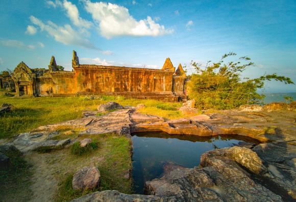 Photo shows an old temple seen from the south. It is placed behind an artificial small basin of water that mirrors the lightly clouded blue sky. The area with the basin is a stony area with large blocks. Behind it, there is a green turf area where the temple is situated. Behind the temple other buildings are visible, as is some clusters of trees. It is closing in on afternoon and sunset, and the sunlight is warm and yellow giving extra deep colours to the flora, stones and temple.