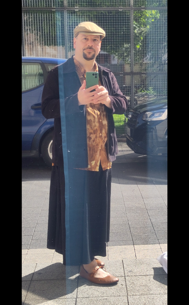 Selbstporträt in spiegelnder Scheibe. Ich trage einen langen schwarzen weiten Rock, ein braun-beige unregelmäßig gemustertes Hemd, braune Ballerinas mit Schnürung, eine schwarze Jacke und hellbraune Schiebermütze aus Stoff.