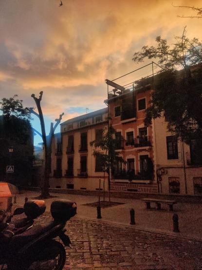 Edificios al atardecer en la plaza de Santo Domingo. En el cielo se ven unas nubes naranjas, iluminadas por los últimos rayos del sol. Delante de los edificios hay unos árboles.
