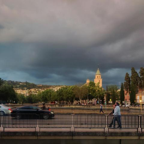 Una pareja camina por el puente del Paseo  del Violón. Al fondo se ve el puente Romano y más allá la torre de los Escolapios iluminada por los últimos rayos del sol. Hay unas nubes grises en el cielo que le dan a la escena un tono dramático.