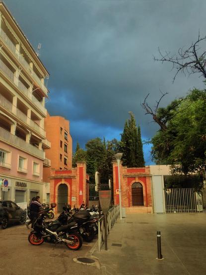 Entrada al Cuarto Real de Santo Domingo. Justo al lado hay un edificio cuya entrada es de color rojo. Hay un aparcamiento de motos justo delante, lleno hasta arriba. Al fondo se ven unas nubes oscuras que amenazan lluvia.