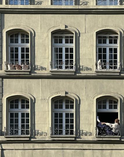 An array of six windows—two floors, with three each—on the side of an old stone building whose wall is painted a greyish green. The windows are arched and have small wrought-iron Juliet balconies. The bottom-right one is open and someone is lounging in the balcony.