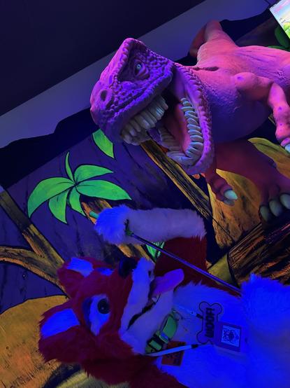 A red and white doggo fursuiter underneath a black light posing with a dino statue at a putt putt golf course.