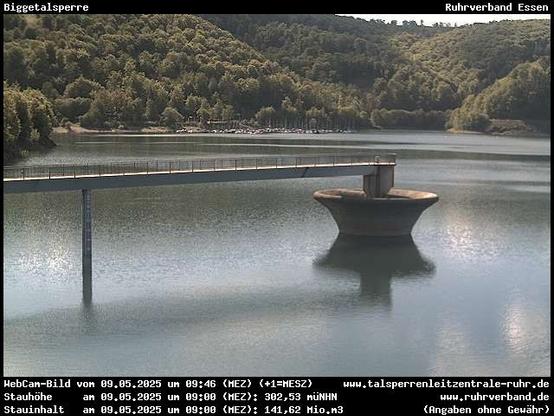 Das Bild zeigt die Wasserfläche der Biggetalsperre. Von links ragt eine schmale Brücke ins Bild, die an der trichterförmigen Hochwasserentlastungsanlage endet. Im Hintergrund ist ein Bootshafen, darüber ein bewaldeter Hang zu sehen.