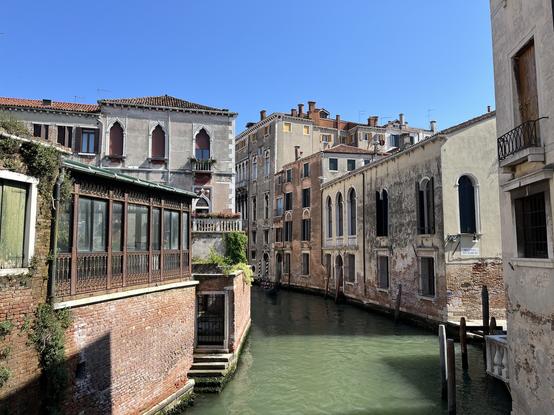 Eine ruhige Kanalszene in Venedig, die verwitterte Gebäude mit gewölbten Fenstern, einem klaren blauen Himmel und Reflexionen im Wasser zeigt.