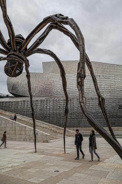 A portrait view taken outside a building with a modern architectural design on an overcast day. The building in the background is shaped as if great sheets of metal were wrapped and bent around one another, all of it encased in smaller metal panels that look a little like the scales of some metallic beast from this distance. It's a combination of harsh materials and some flowing areas and still come up to sharp edges and points in places. In the foreground this is an open area paved with pale flagstones and there is a wide set of stone steps leading up to the left away from the building behind. A couple of people are on the steps and there are three more walking across this space in front with two of those passing between the legs of a sculpture that dominates the near view. The sculpture is a spider in form with legs that are long, thin cables or muscles stretching up about ten metres to then arch down a little to the central body of the piece, two spherical cage constructions atop one another, one above, one below the level where the legs join. Within the caged lower part there are metal eggs. The sculpture is a nightmare vision of an arachnid.