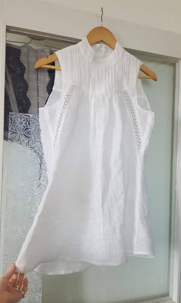 A white sleeveless top hanging off a clothes hanger which in turn is hanging off a glass door. It has a closure in the back yoke, the front yoke is heavily decorated with little tucks, pintucks and lace. From the yoke falls a voluminous skirt section, to roughly mid hip on a person. There is a bit of diagonal insertion lace on the upper front and side, looking very intentional and not at all something to piece together a cutting mistake! Sini's hand is holding onto the hem a bit. Despite the amount of fabric gathered onto the yoke the linen falls and smooths down gracefully, and would hug the body of the wearer loosely, for it is just so soft. The fabric is sheer. Behind the door is a gray corset with black lace, just hanging out.