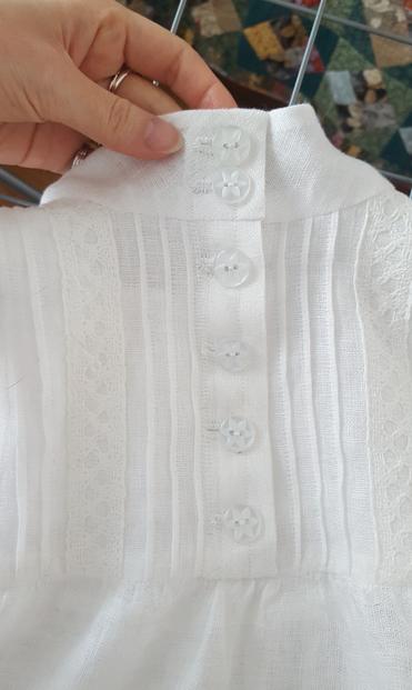 Detail photo of the back button placket. It's only the length of the yoke, which stops at about shoulder blade level. There are four buttons on the body and two on the collar, which is a medium to tall standing collar. There are more pintucks and more lace. The buttons themselves are a 1.5 centimetre white pearlescent plastic, with a flower motif, easier to button with arthritic fingers than a tiny button. They sit in hand sewn buttonholes. The fabric is so soft and sheer you can see through the doubled up yoke fabric as well. Sini's pink fingers hold onto the collar, a bit of quilt in the background.