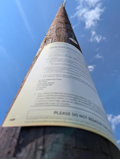 Wide-angle, colour photograph of a wooden telegraph pole, looking up. The exaggerated perspective draws the pole to a sharp point at the top of the picture. No wires have been attached yet; the pole is brand new, and still bears its installation notice, which has been stapled to the wood.