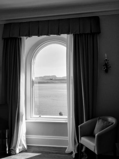 A rounded window against the light. Behind it you can see a stretch of water and mountains in the distance. The window is anchored with curtains and open shades. A faux-candle sconce is on the right-hand wall. A club armchair with a cushion is next to it.