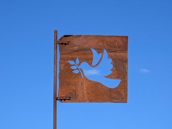 Das Bild zeigt eine rostige Metallplatte, die wie eine Fahne an einer Metallstange befestigt ist. Aus der Platte ist die Silhouette einer Taube mit einem Olivenzweig im Schnabel ausgeschnitten. Durch diesen Ausschnitt ist der klare blaue Himmel mit einer kleinen, vereinzelten Wolke im Hintergrund sichtbar. Die Metallplatte und die Stange weisen deutliche Rostspuren auf, was auf Alter und Witterungseinflüsse hindeutet. Die Kanten der Platte sind teilweise unregelmäßig und ausgefranst.