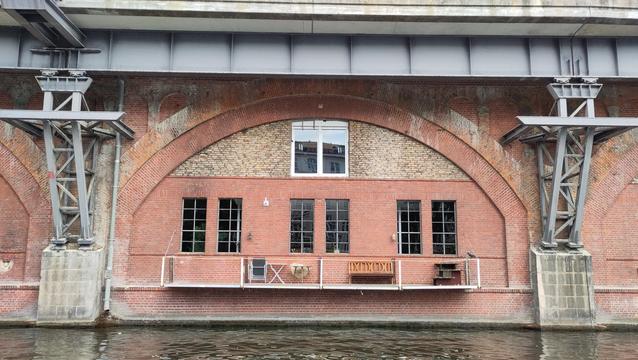 eine rote Klinkerwand, in der oberen Hälfte mit einem halbkreisförmigen Bogen aus ebenfalls roten Klinkern, darin oben ein zweiflügeliges Fenster mit weißem Rahmen, in der unteren breiteren Hälfte drei Fensterpaare mit Sprossenfenstern, die Rahmen dünner und hellgrau; vor diesen sechs Fenstern ist ein filigraner Balkon mit dünnem grauem Geländer montiert, darauf stehen von links nach rechts ein zusammengeklappter Stuhl, ein aufgestellter Klapptisch, ein dreibeiniger Tisch, eine Holzbank und rechts eine zusammengezimmerte Treppe, um aus dem rechten Fenster auf den Balkon zu gelangen; rechts auf dem Geländer ist eine Krähenattrappe montiert; links und rechts am Bildrand massive graue Sandsteinpfeiler, auf denen stählerne Gitterstützen an der Wand nach oben ragen, sie tragen einen breiten grauen Stahlträger, der quer über den oberen Bildrand verläuft und eine Ebene aus Beton trägt, vmtl. eine Brücke. Am unteren Bildrand Wasser entlang der Mauer