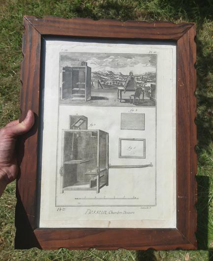 A hand holding an engraving in a wooden frame. The engraving is entitled 'Le Dessein' and shows different types of portable camerae obscurae, meant for tracing the views inside of them