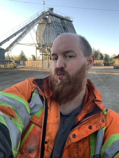 A cis white male offer a hug. He has a red full beard and wears a reflective jacket. Behind him the sun is coming up
