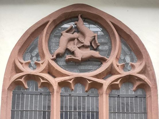 Oberer Teil eines Kirchenfensters von außen, in der Rosette sind aus Stein drei springende Hasen im Kreis so angeordnet, dass sie die Ohren gemeinsam haben, also drei Hasen mit jeweils 2 Ohren aber insgesamt nur 3 Ohren.