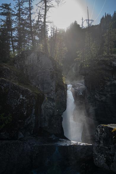Looking toward the setting sun. A wall of rock is surmounted by trees, pierced by the cleft that allows water to flow through and over the falls, creating a lot of mist. The sun shining through the cleft lights up the mist.