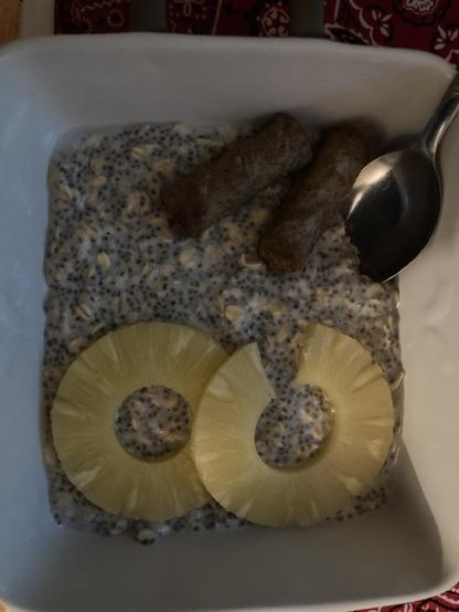 A dish of oatmeal, with chia seeds, two pineapple slices, and two link sausages