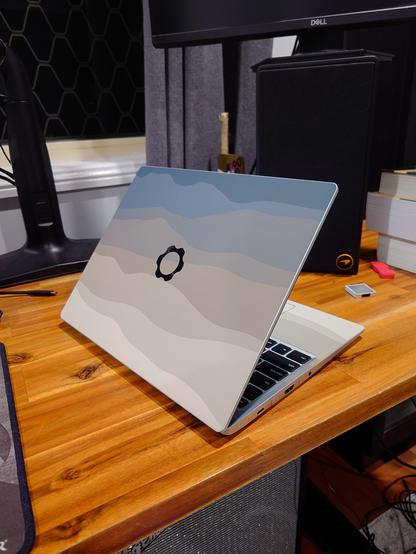 A Framework Laptop 13 sitting on a wooden desk, with the Dbrand Alpine skin applied on the lid, trackpad and deck. A USB-C and HDMI expansion card are inserted, and a DisplayPort card is sitting on the desk in front of a speaker.