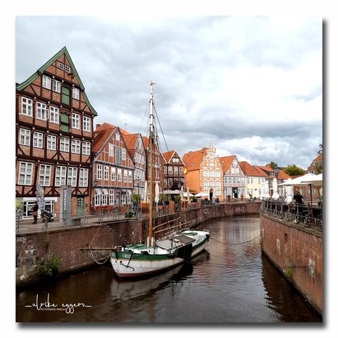 Alte, zum Teil schiefe Fachwerkhäuser im alten Hansehafen von Stade. Im Vordergrund liegt ein kleines, weiß-grünes Segelschiff im Wasser. Im Hintergrund befinden sich weitere Gebäude und einige Menschen sitzen an den Außenterrassen von Restaurants oder Cafés. Der Himmel ist bewölkt.