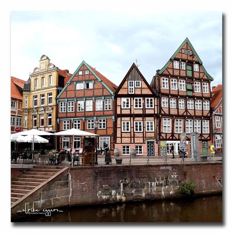 Noch einmal der Hansehafen von Stade mit den alten, schiefen Fachwerkhäusern. Vor den Gebäuden befindet sich ein Bereich mit von großen Schirmen überdachten Tischen und Stühlen, der zu einem Café gehört. Am linken Bildrand führt eine Treppe aus Ziegelsteinen zum Fluß hinunter.