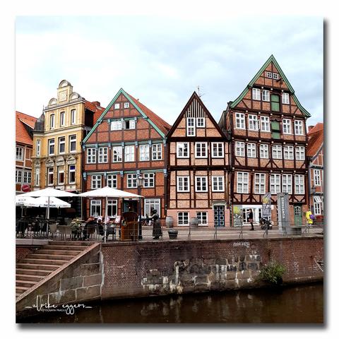 Noch einmal der Hansehafen von Stade mit den alten, schiefen Fachwerkhäusern. Vor den Gebäuden befindet sich ein Bereich mit von großen Schirmen überdachten Tischen und Stühlen, der zu einem Café gehört. Am linken Bildrand führt eine Treppe aus Ziegelsteinen zum Fluß hinunter.