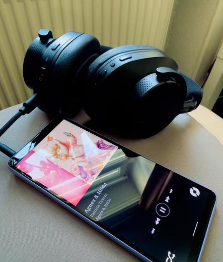 Screen on a pillow displaying the music cover of Patricia Taxxon's album Agnes and Hilda and player controls. Black over ear headphones behind it.