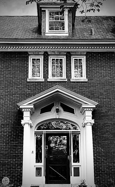 Image of a 1920s Georgian Revival brick house. Rust Belt Viewpoint-2025- Georgian Revival residential home ca. 1925, Broad Ripple, Indianapolis, Indiana, USA. Image shows a typical Georgian Revival Colonial home, with a columned front port surmounted by a detailed gabled entablature with a stylized transom window, above, three windows, two two over three, the center three over three paned, surmounted above a detailed dentil moulding by a roof dormer with double-hung window with two three-over-two panels.