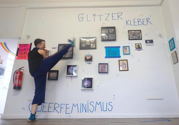Alex presenting her art on the wall with a high-kick. There are several picture-frames with collages in them.