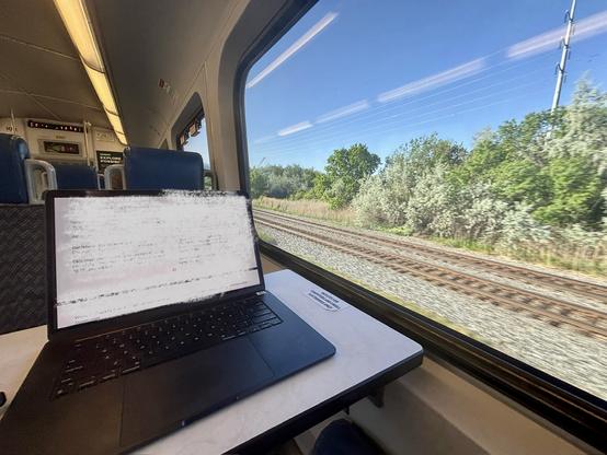 A laptop sits on a table next to a window. Outside, trees and railroad tracks pass by.